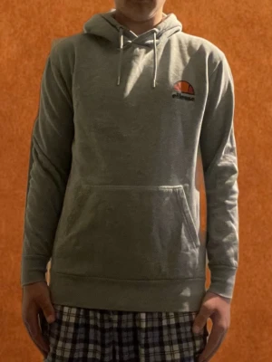 Grå Ellesse hoodie med logga - Säljer en grå hoodie från Ellesse med klassisk logga på bröstet. Tröjan har huva med dragsnören och en stor magficka. Materialet är mjukt och bekvämt, perfekt för chill dagar. Långa ärmar och ribbade muddar ger en snygg passform. Mitt pris 200 skriv vid funderingar 