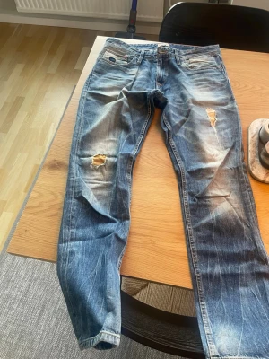 Jeans Hilfiger Denim - Säljer ett par blå jeans från Hilfiger Denim med slitningar och hål på benen. Jeansen har klassisk femficksdesign, knappgylf och är tillverkade i denim. Perfekt för dig som gillar en cool och avslappnad look.