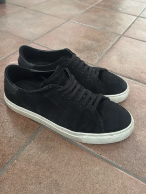 Svarta Axel Arigato sneakers i mocka - Stilrena svarta sneakers i mocka med matchande svarta skosnören och vit sula. Låg modell med enkel design som passar till det mesta. Skorna har en klassisk siluett och är perfekta för dig som gillar en clean look.                                                                                 En sömm har öpnnats lite på den högra skon men kan lagas med skolim eller hos en skomakare. Låda och dustbag ingår. Pris kan diskuteras 