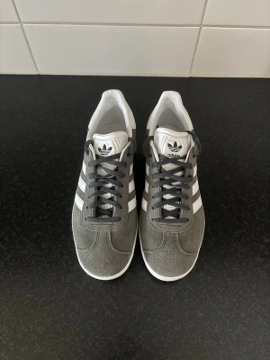 Adidas Gazelle grå sneakers - Snygga Adidas Gazelle sneakers i grå mocka med vita detaljer och klassiska tre ränder på sidan. Skorna har mörkgrå snörning, vit sula och vit häl. Perfekt för dig som vill ha en ikonisk och stilren sneaker med retrovibe.