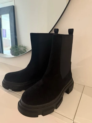 Chunky svarta Chelsea boots - Säljer ett par svarta Chelsea boots med chunky sula och klassisk resår på sidorna. Bootsens ovandel är i mockaliknande material och de har dragflikar både fram och bak. Perfekta för dig som vill ha en edgy och stilren look.