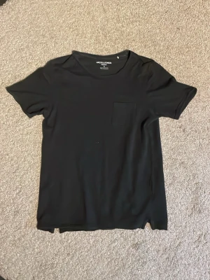 Svart t-shirt från Jack & Jones - En enkel svart t-shirt från Jack & Jones med rund hals och liten bröstficka. T-shirten har korta ärmar och är gjord i mjuk bomull, perfekt för en avslappnad och stilren look. Stl 38 tog jag bara för att kunna lägga upp annonsen så vet ej om det är rätt, men annars är stl S