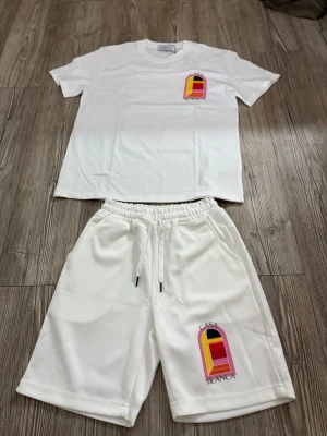 Vitt set från Casablanca med tryck - Säljer ett vitt set från Casablanca med färgglatt grafiskt tryck på både t-shirt och shorts. T-shirten har rund hals och korta ärmar, shortsen har resårmidja med dragsko och sidofickor. Materialet ser ut att vara mjukt och bekvämt, perfekt för en avslappnad stil.