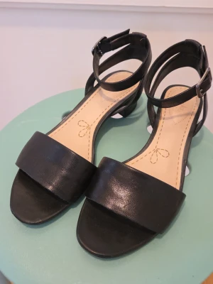 Svarta sandaler med ankelrem - Stilrena svarta sandaler från Clarks med bred rem över tårna och justerbar ankelrem med spänne. Sandalerna har låg klack och en mjuk innersula i beige med dekorativ söm. Perfekta för en trendig och bekväm look.