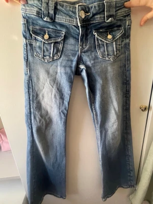 Blå bootcut jeans med fickdetaljer - Snygga blå bootcut jeans med slitningar och coola fickdetaljer framtill. Jeansen har breda hällor och dekorativa knappar på fickorna. Klassisk femficksmodell i denim med ljus tvätt och markerade sömmar. Perfekta för en avslappnad och trendig look.