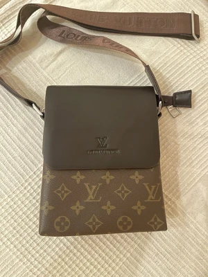  axelväska helt ny  - Snygg axelväska  i klassiskt brunt monogrammönster. Väskan har ett lock i mörkbrunt läder och justerbar axelrem med logga. Perfekt storlek för det viktigaste och en stilren accessoar till din outfit.