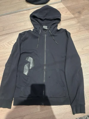 Svart hoodie från Peak Performance - Säljer en svart hoodie från Peak Performance med dragkedja framtill, två fickor och justerbar huva med snören. Diskret logga på bröstet i svart. Tillverkad i mjukt bomullsmaterial som är skönt att ha på sig. Perfekt för en avslappnad och stilren look.