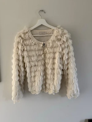 Krämvit fransig kofta från Cream - Unik krämvit kofta från Cream med fransdetaljer över hela plagget. Koftan har rund halsringning, dragkedja framtill och är tillverkad i ett lätt material. Storlek M men passar även s och xs. Bra skick då den kommit till användning fåtalet gånger.