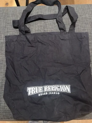 Svart shoppingväska True Religion - Snygg svart shoppingväska från True Religion med vitt tryck på framsidan. Väskan har dubbla handtag och är gjord i ett mjukt tygmaterial. 