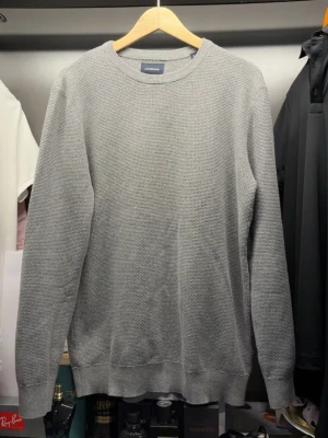 Grå stickad tröja från Lindbergh - En stilren grå stickad tröja från Lindbergh med rund halsringning och diskret strukturmönster. Tröjan har långa ärmar och ribbade muddar vid ärmslut och nederkant. Perfekt för lager-på-lager och enkel att matcha.