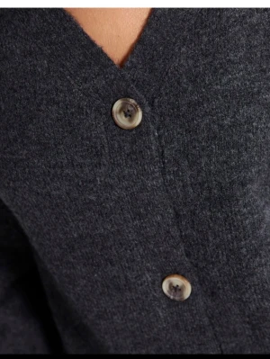 Mörkgrå kofta med knappar - Mysig mörkgrå kofta med v-ringning och stora, marmorerade knappar framtill. Koftan har en avslappnad passform och är tillverkad i ett mjukt, stickat material som passar perfekt till jeans eller kjol.