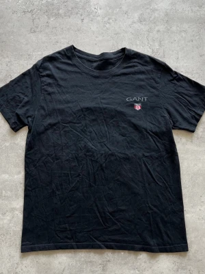 Svart GANT t-shirt med logga - Klassisk svart t-shirt från GANT med rund hals och korta ärmar. På bröstet finns en liten broderad GANT-logga och ett sköldmärke i vitt och rött. Tillverkad i mjuk bomull för en skön känsla.