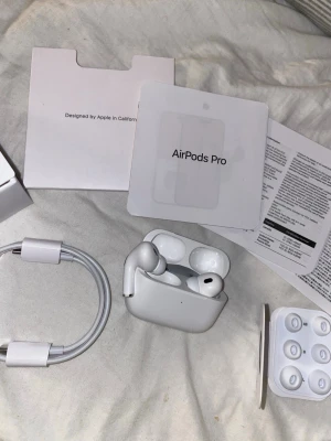 Apple AirPods Pro 2nd Gen hörlurar - Säljer ett par vita Apple AirPods Pro (2nd generation) med laddningsetui, USB-C kabel och extra silikonpluggar. Hörlurarna har en stilren och kompakt design i plast och levereras med originalförpackning och manualer.