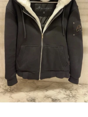 Svart teddyjacka Moose Knuckles - Säljer en svart teddyjacka från Moose Knuckles med vit fuskpäls på insidan. Jackan har dragkedja framtill, två stora fickor och en logga på ärmen. Perfekt för kalla dagar och har en skön, oversized passform.