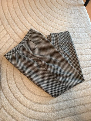 Rutiga kostymbyxor i svart och beige  - Snygga kostymbyxor med klassiskt rutigt mönster i svart och beige . Byxorna har rak passform och pressveck, vilket ger en stilren look. Perfekta att matcha med en enkel topp eller skjorta. Materialet är  mjukt och följsamt.