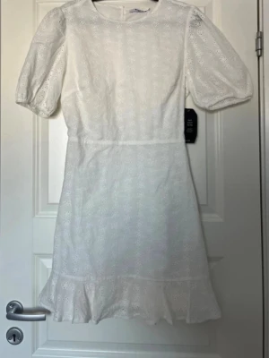 Vit broderad kortklänning från NA-KD - Söt vit kortklänning från NA-KD med broderade blommor, puffärmar och volangkant nertill. Klänningen har rund halsringning och är tillverkad i bomull, vilket gör den både luftig och bekväm. Perfekt för sommarens alla tillfällen!