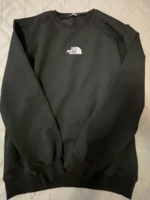 Svart sweatshirt från The North Face - Klassisk svart sweatshirt från The North Face med rund halsringning och vit logga broderad på bröstet. Tröjan har ribbade muddar vid ärmslut och nederkant, perfekt för en clean och enkel stil.