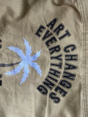 Grön overshirt med tryck och fickor - Snygg grön overshirt med svart texttryck 'Art changes everything' och blå palm på ryggen. Modellen har två bröstfickor med lock och knappar framtill. Perfekt att slänga över en t-shirt eller klänning för en chill look. Från Zadig et Voltaire o psssar ca S❤️