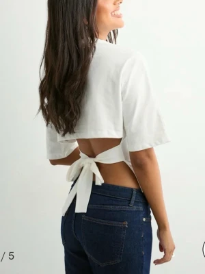 Vit croppad topp med knyt i ryggen - Trendig vit croppad topp från Nelly med halvlånga ärmar och snygg knytning i ryggen. Toppen har en enkel, stilren framsida och öppen rygg som ger en modern touch. Perfekt för dig som vill ha en unik och fräsch look.