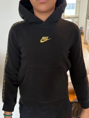 Svart Nike hoodie med gult tryck - Svart hoodie från Nike med gul logga på bröstet och gul Nike-text längs ärmarna. Klassisk känguruficka framtill och huva. Tillverkad i mjukt material som passar perfekt till en avslappnad streetwear-look.