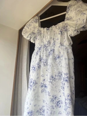 Vit blommig kortklänning med volang - Söt vit kortklänning med blått blommönster och volangdetaljer vid axlarna. Klänningen har en smockad rygg och puffiga korta ärmar. Perfekt för sommarens alla tillfällen och ger en romantisk vibe.