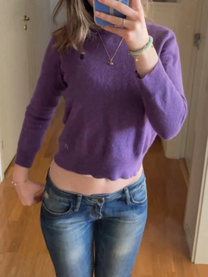 Lila stickad tröja Polo Ralph Lauren - Lila stickad tröja från Polo Ralph Lauren med rund halsringning och diskret broderad logga på bröstet. Tröjan har lång ärm och en något croppad passform med vågig kant nedtill. Perfekt för dig som gillar en färgglad och trendig look.