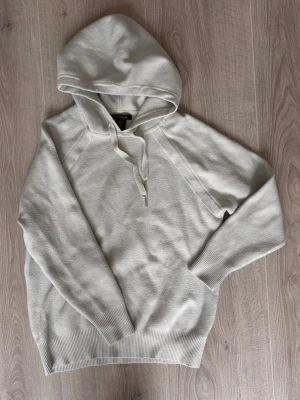 Vit hoodie från Tahari med huva - Säljer en stilren vit hoodie från Tahari i storlek M. Hoodien har en klassisk huva med dragsko och ribbade muddar vid ärmslut och nederkant. Perfekt för dig som gillar en enkel och clean look. Materialet känns mjukt och bekvämt mot huden.