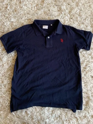 Mörkblå pikétröja från US Polo Assn  - Mörkblå pikétröja från US POLO med klassisk krage, korta ärmar och röd broderad logga på bröstet. Tröjan är gjord i bomull och har en stilren och tidlös design med knappslå framtill. Storlek”12-13 år” (146-152) Pris kan diskuteras!