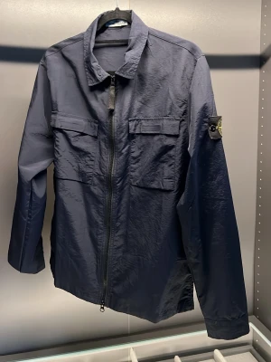 Mörkblå overshirt från Stone Island - Snygg mörkblå overshirt från Stone Island med dragkedja framtill, två bröstfickor med lock och den klassiska logotypen på vänster ärm. Tillverkad i ett lätt, glansigt material som ger en modern look. Perfekt lager-på-lager-plagg.