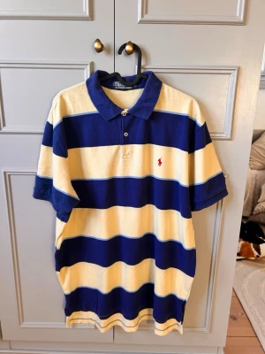 Randig pikétröja från Polo Ralph Lauren - Klassisk pikétröja med breda blå och ljusgula ränder från Polo Ralph Lauren. Tröjan har krage, två knappar vid halsen och den ikoniska röda logotypen broderad på bröstet. Tillverkad i mjuk bomull som känns skön mot huden. Srl (L)