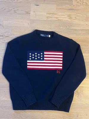 Mörkblå stickad tröja Polo Ralph Lauren - Mörkblå stickad tröja från Polo Ralph Lauren med amerikansk flagga i rött, vitt och blått på bröstet samt RL-broderi. Tröjan har rund halsringning och ribbade muddar vid ärmslut och nederkant. Perfekt för dig som gillar klassisk college-stil.