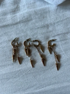Guldiga örhängen med kedjor och spikes - Säljer fyra par coola örhängen i guldfärgad metall med små kedjor och hängande spikes. Örhängena har en edgy vibe och passar perfekt för dig som gillar statement-accessoarer. Ingen synlig märkning på örhängena.