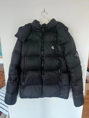 Svart Calvin Klein jacka - Snygg svart pufferjacka från Calvin Klein med avtagbar luva och dragkedja framtill. Jackan har två stora fickor med dragkedja och en innerficka och CK-logga på bröstet och ärmen. Bra för vår och höstdagar men lite för tunn för vintern.