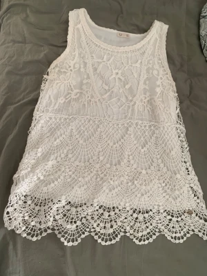 Vit spetslinne från Ü - Supersött vitt linne från Ü med detaljerad spets och broderade mönster. Ärmlös modell med rund halsringning och vacker scallop-kant nedtill. Perfekt för sommarens alla tillfällen och ger en romantisk vibe till din outfit.