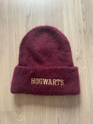 Vinröd Hogwarts mössa - Säljer en vinröd stickad mössa med uppvikt kant och gul brodyr med texten 'HOGWARTS' framtill. Perfekt för dig som älskar Harry Potter och vill hålla dig varm med stil. Mössan har en klassisk form och är gjord i mjukt material.