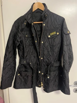 Svart quiltad jacka från Barbour - Snygg svart quiltad jacka från Barbour med bälte i midjan, gulddetaljer och flera fickor. Jackan har hög krage, dragkedja och knappar framtill samt Barbour-logga på bröstet. Perfekt för dig som gillar klassisk och tidlös stil.
