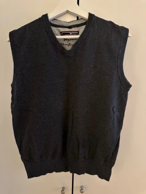 Mörkgrå stickad väst med v-ringning - Klassisk mörkgrå stickad väst med v-ringad hals och ribbade kanter. Perfekt att bära över en skjorta för en stilren look. Enkel och tidlös design utan mönster eller extra detaljer.