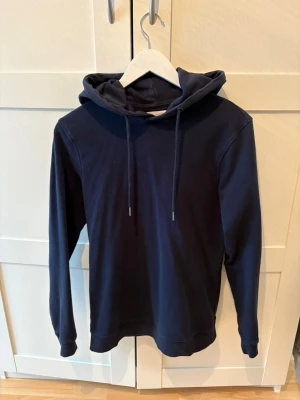 Mörkblå hoodie  - En stilren mörkblå hoodie med huva och dragsnören. Hoodien har långa ärmar och ribbade muddar vid ärmslut och nederkant. Tillverkad i mjukt bomullsliknande material som känns skönt mot huden. Perfekt för en avslappnad look.
