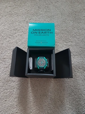 Omega x Swatch Polar Lights klocka - Säljer en Omega x Swatch Mission on Earth Polar Lights klocka med turkos boett och svart urtavla. Klockan har kronograf, tydliga index och svart textilband med Omega-logga. Kommer i originalbox och är en del av Bioceramic MoonSwatch-kollektionen.