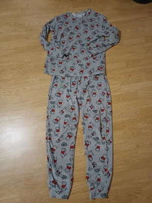 Grå Disney Nalle Puh pyjamas - Mysig grå pyjamas med långärmad tröja och byxor, täckt av ett gulligt Nalle Puh-motiv i rött och svart. Perfekt för chillkvällar hemma. Mjukt material som känns skönt mot huden. Disney-logga tryckt på flera ställen.