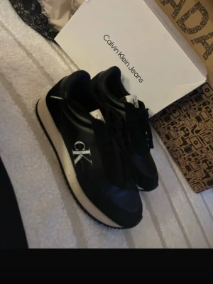 Svarta Calvin Klein Jeans sneakers - Stilrena svarta sneakers från Calvin Klein Jeans med vit sula och stort CK-logotyp på sidan. Skorna har klassisk låg profil, snörning och ovandel i syntetmaterial och textil. Perfekta för dig som gillar en clean och modern look. Mocha och skin material 