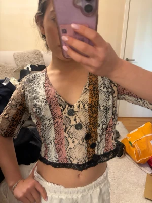 Croppad blus med ormmönster och knappar - Säljer en croppad blus med v-ringning och korta ärmar. Blusen har ormmönster i beige, rosa och bruna toner samt svarta knappar framtill. Nedtill finns en svart spetskant.