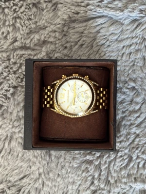 Guldig Michael Kors klocka med stenar - Snygg guldig klocka från Michael Kors med vit urtavla och gnistrande stenar runt boetten. Klockan har länkarmband i metall, flera små urtavlor och tydliga guldfärgade detaljer. Perfekt accessoar för att lyfta din stil.
