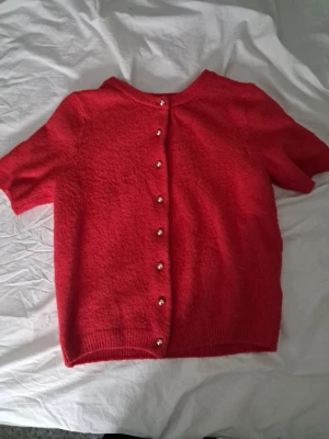 Röd stickad topp med pärlknappar - Snygg röd stickad topp med korta ärmar och dekorativa pärlknappar längs ryggen. Tröjan har rund halsringning och ribbade kanter. Perfekt för dig som vill ha något unikt och trendigt i garderoben.