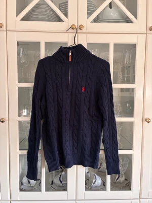 Mörkblå kabelstickad tröja Polo Ralph Lauren - Kabelstickad mörkblå tröja från Polo Ralph Lauren med halv dragkedja och hög krage. Tröjan har det klassiska röda Polo-logot broderat på bröstet och ribbstickade muddar vid ärmslut och nederkant. Perfekt för lager på lager och snygg till jeans.