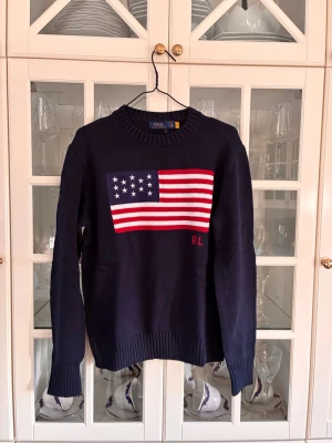 Mörkblå stickad tröja Polo Ralph Lauren - Säljer en mörkblå stickad tröja från Polo Ralph Lauren med amerikansk flagga i rött, vitt och blått på bröstet samt RL-broderi. Tröjan har rund halsringning och ribbstickade muddar vid ärmslut och nederkant. Perfekt för dig som gillar klassisk preppy stil.