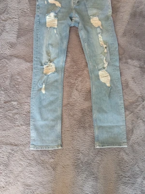 Ljusblå slitna  jeans - Säljer ett par ljusblå jeans med flera slitningar och hål framtill samt på bakfickorna. Jeansen har klassisk femficksmodell och är tillverkade i denim med stretch för skön passform. Perfekta för en trendig och avslappnad look.