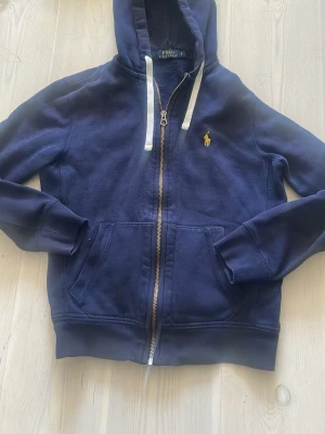 Marinblå hoodie från Polo Ralph Lauren - Snygg marinblå hoodie från Polo Ralph Lauren med dragkedja, vit dragsko och klassisk gul logga på bröstet. Hoodien har kängurufickor och ribbade muddar. Perfekt för en avslappnad och stilren look.