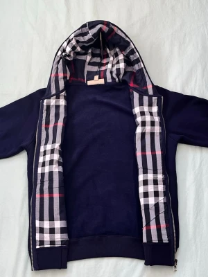 Marinblå hoodie från Burberry - Snygg marinblå hoodie från Burberry med dragkedja och klassiskt rutigt foder i huvan och längs insidan. Hoodien har två fickor fram, ribbade muddar och en diskret Burberry-logga på bröstet. Perfekt för dig som gillar stilrena detaljer och exklusiv känsla.