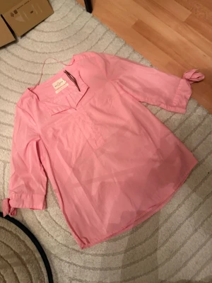 Rosa blus från Holly & White - Snygg rosa blus från Holly & White med v-ringad hals och trekvartsärmar som kan knytas vid ärmsluten. Tillverkad i ett tunt, lätt material som känns luftigt och bekvämt. Perfekt att styla med jeans eller kjol för en fräsch look.